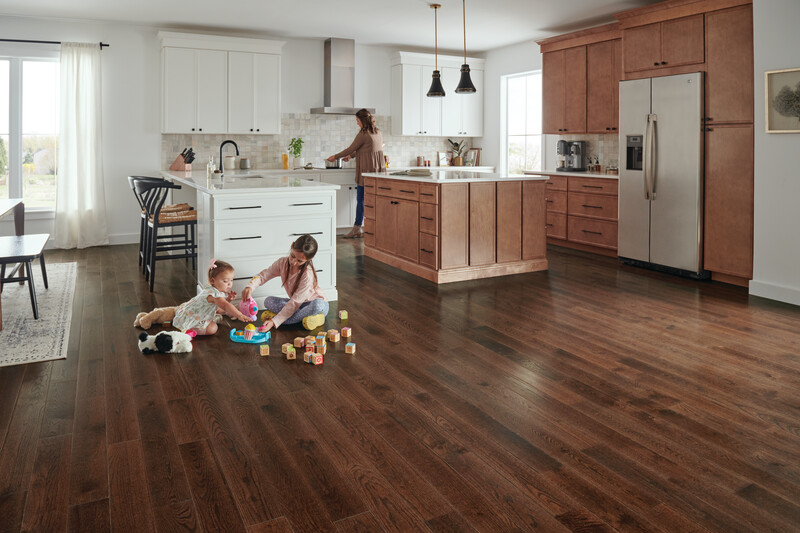 An infant and her sister playing on a hardwood floor in the kitchen with their mother placing a pot on the stove